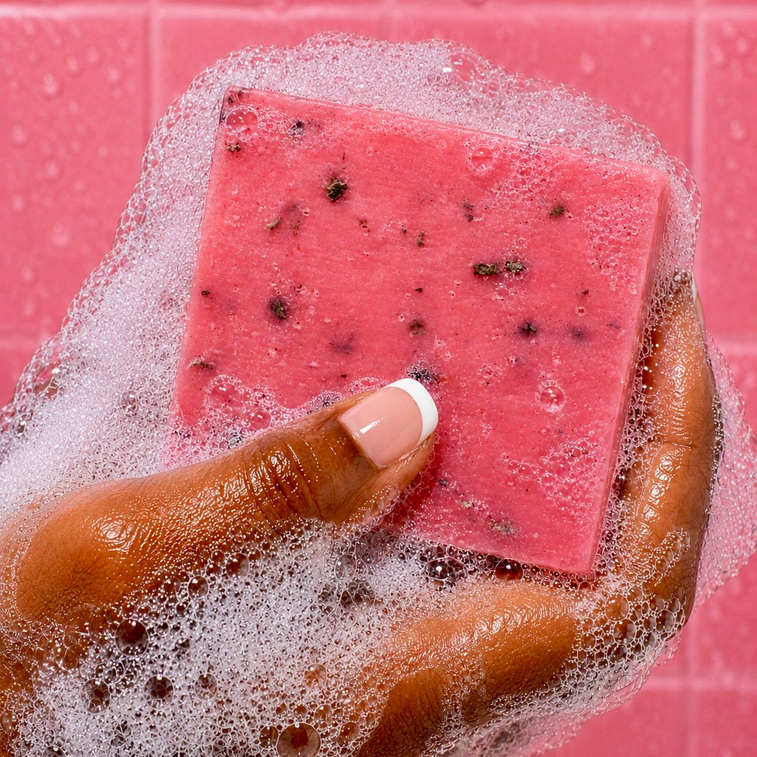 Hand holding a Watermelon soap bar of soap with black specks against a pink tiled background